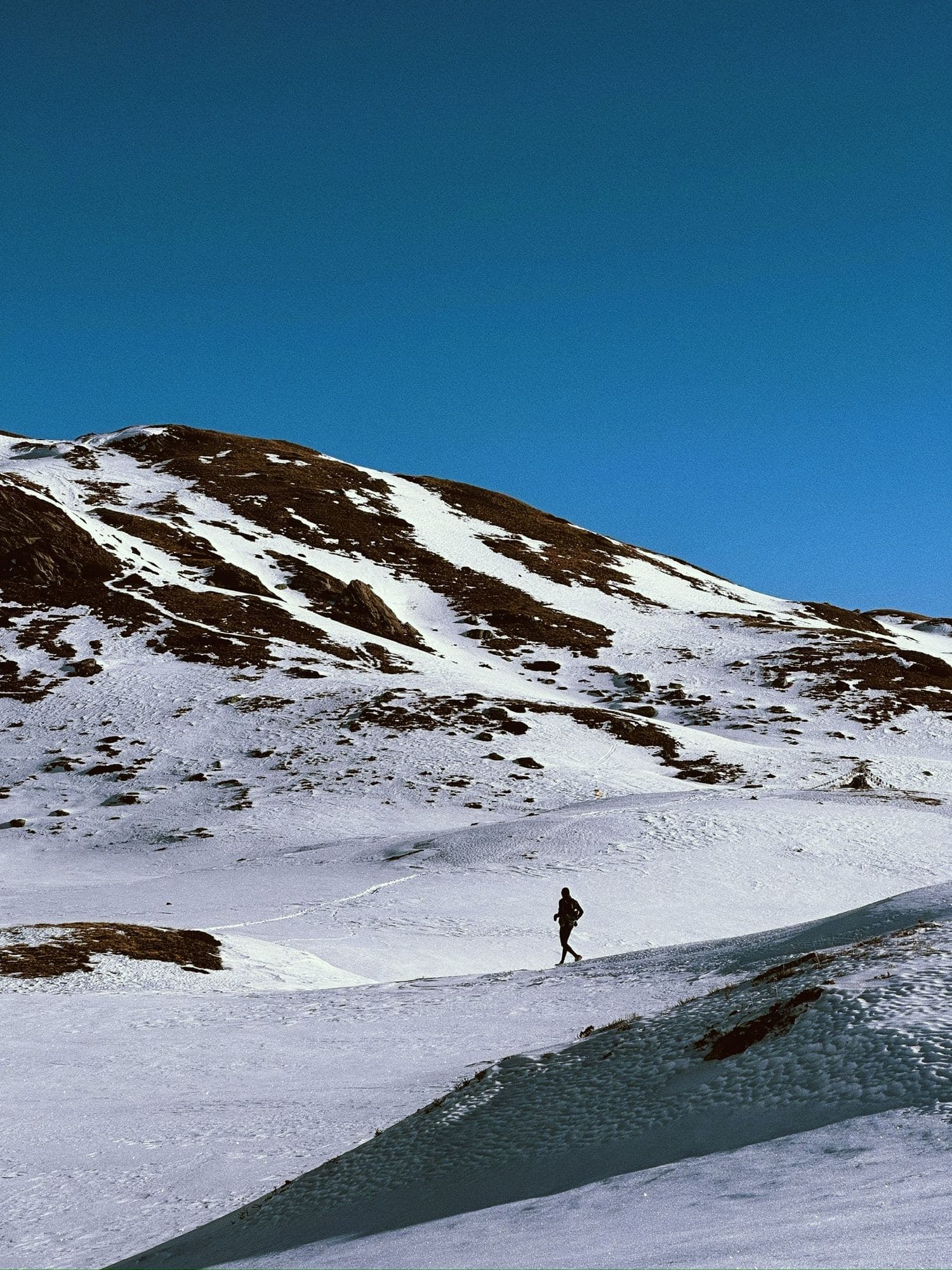 A runner crossing a snowfield below a mountain under a deep blue sky