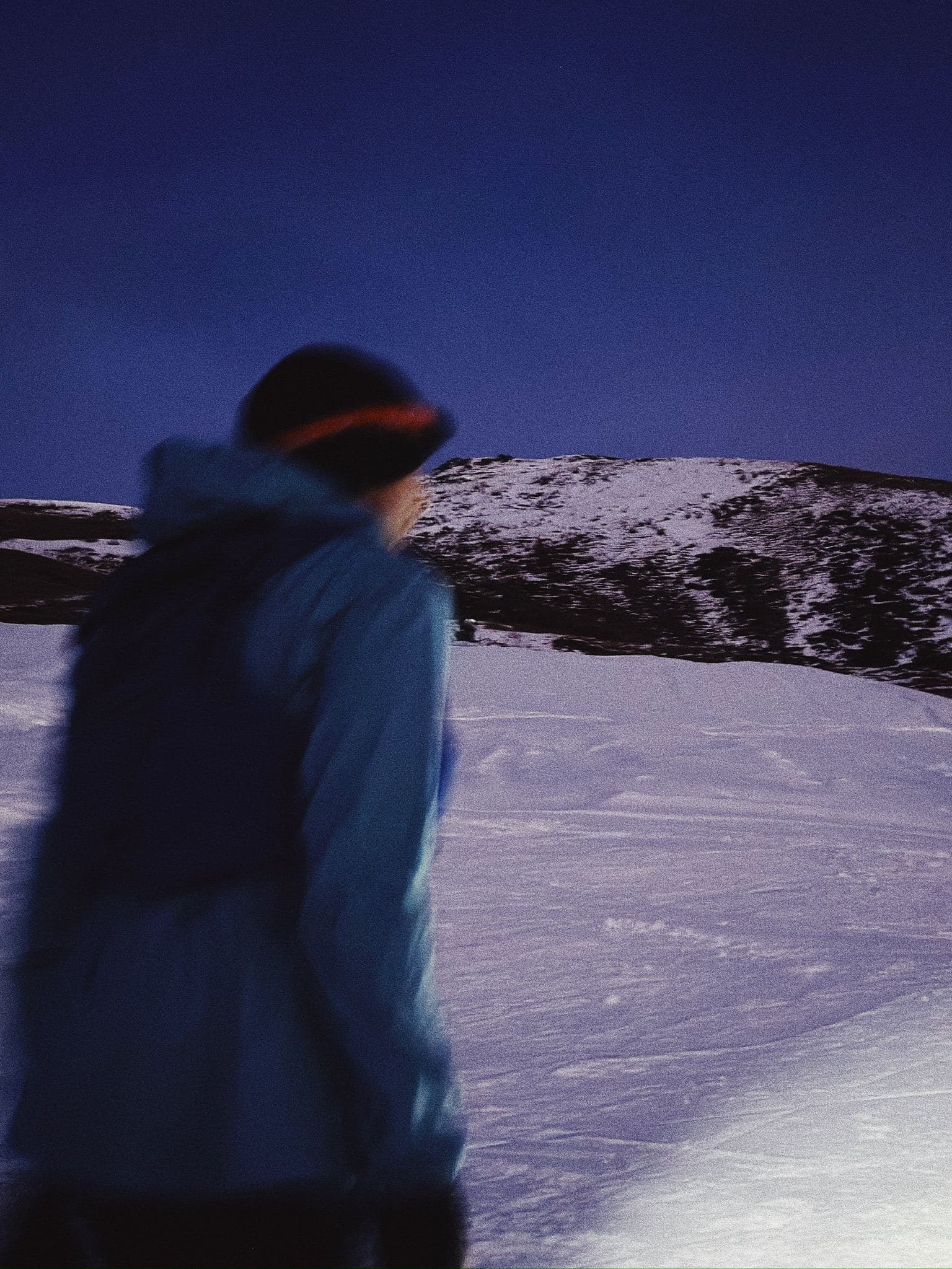 A runner in a winter jacket and headlamp crossing a snowfield at dusk